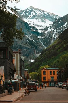 the mountains are covered in snow as people walk down the street near buildings and parked cars