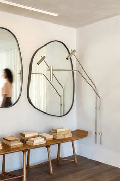 two mirrors on the wall above a table with books in front of it and a woman walking past them