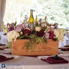 a wooden box with flowers and wine bottles in it on top of a white table cloth