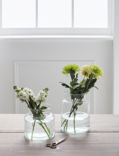 two clear vases with flowers in them on a wooden table next to a pair of scissors