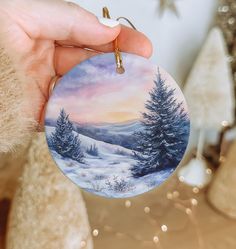 a hand holding a christmas ornament with a snowy landscape on it and trees in the background