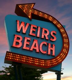 a sign that says weirs beach with an arrow pointing to the right in front of a sunset sky