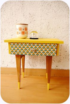 a small yellow table with drawers and a cup on it's top, next to a white wall