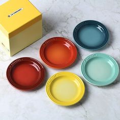 four different colored plates sitting next to a box on a marble counter top with the lid open