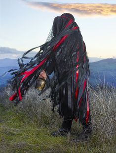 a person standing on top of a grass covered hill with long black hair in the wind