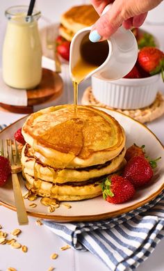 pancakes with syrup being poured onto them and strawberries on the plate next to it