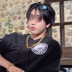 a young man wearing a black shirt and gold chain around his neck, sitting in front of a stone wall