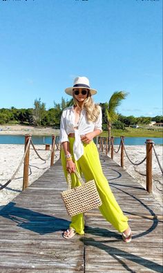 a woman in yellow pants and hat standing on a dock with a straw bag over her shoulder