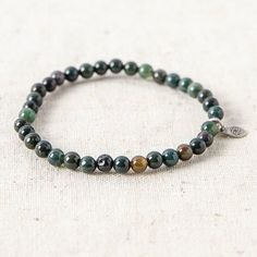 the bracelet is made with green and brown beads on a white cloth background, it has a silver charm