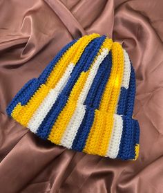 a crocheted hat laying on top of a brown satin covered bed sheet with it's yellow, white, and blue stripes