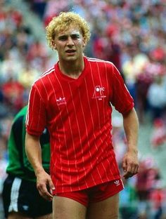 a man in red shirt and shorts on soccer field with crowd watching from the sidelines