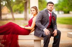 a man and woman sitting on a bench in front of some trees wearing formal clothes