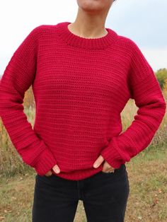 a woman wearing a red sweater standing in front of a field with her hands on her hips