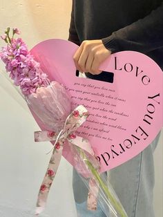 a person holding a pink heart shaped sign with flowers in it and a message written on it