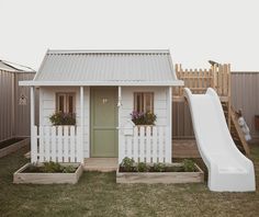 a small white house with a slide and flower boxes in the front yard, next to a wooden fence