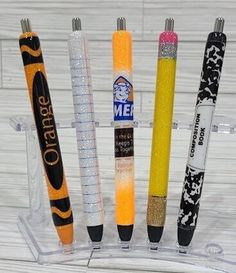 four pens are in a clear holder on a white wooden surface, one is yellow and the other is black