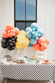 a table topped with lots of balloons next to a white wall and window covered in checkered cloth