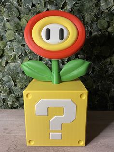 a yellow box with a red flower and question mark on it sitting in front of a green bush