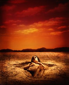 a woman laying on top of a sandy beach under a red sky with clouds in the background