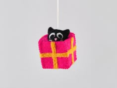 a black cat in a pink box ornament hanging from a white string on a gray background