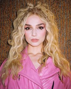 a woman with long blonde hair wearing a pink leather jacket and posing for a photo