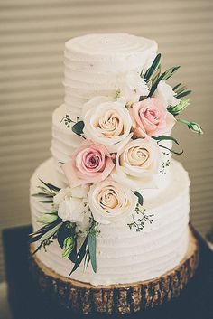 a white wedding cake with pink roses and greenery on the top is sitting on a tree stump