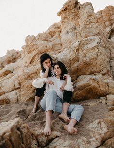 two women sitting on rocks in the sand with their arms around each other and smiling