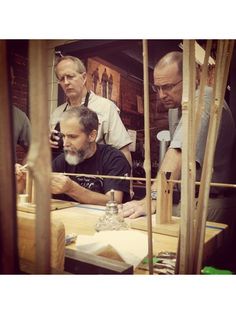 men working on woodworking in a shop with other people looking at the workbench