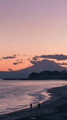 two people walking on the beach at sunset with a mountain range in the distance behind them