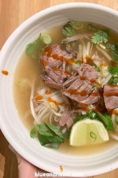 someone holding a bowl of soup with meat, noodles and cilantro on the side