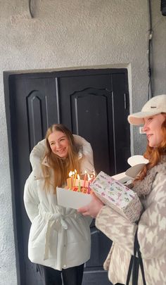 two women standing in front of a door holding a birthday cake