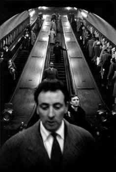 a man wearing a suit and tie standing in front of an escalator filled with people