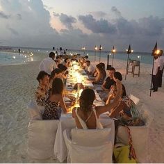 a group of people sitting at a long table on the beach with candles in front of them