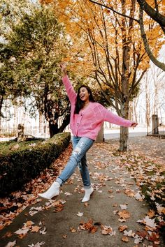 a woman in pink jacket and jeans dancing on sidewalk surrounded by trees with leaves around her