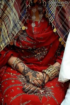 a woman in red sitting down with her hands on her chest and arms covered by jewelry