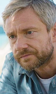 a man with grey hair and beard sitting on the beach looking off into the distance