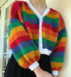 a woman wearing a multicolored cardigan standing next to a white wall and window