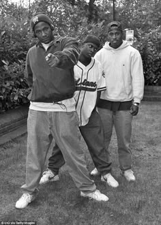 three young men standing next to each other in front of trees and bushes, one pointing at the camera