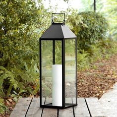 a lit candle sitting on top of a wooden table in front of some bushes and trees