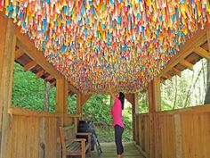 a woman standing under a colorfully decorated ceiling in a wooden structure surrounded by greenery