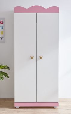 a white and pink cabinet with two gold crowns on the top, next to a potted plant