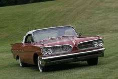 an old red and white car parked on the grass in front of a grassy field