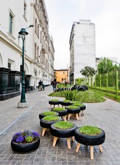 several black tires are lined up on the sidewalk with plants growing out of them and people walking in the background