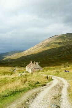 an old stone house in the middle of a grassy field with a dirt road leading to it