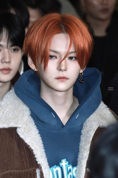 an orange haired man with red hair wearing a blue hoodie and earrings