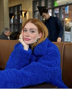 a woman sitting at a table with her hand on her chin, wearing a blue coat