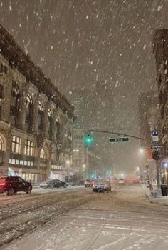 a snowy city street filled with lots of traffic next to tall buildings and green traffic lights