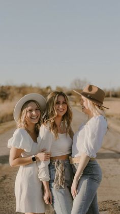 three young women standing together in the middle of an empty road wearing hats and jeans