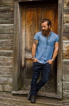 a man with a beard standing in front of a wooden door wearing blue shirt and jeans