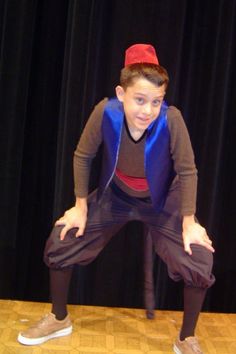 a young boy wearing a red hat and blue vest standing on a wooden floor in front of a black curtain